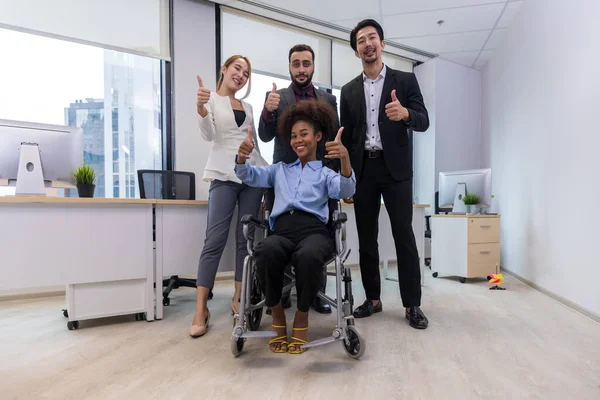 Office workers and woman on a wheelchair in bright office. They are showing a teamwork. Portrait of diverse business team with young woman in wheelchair all smiling at camera in office