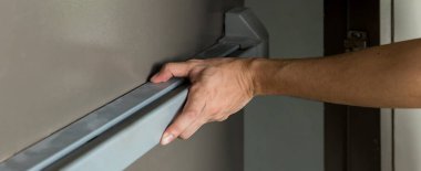 Hand is Opening the emergency fire exit door. Emergency fire exit door. Closed up latch and rusty door handle of emergency exit. Push bar and rail for panic exit. Open one way door.