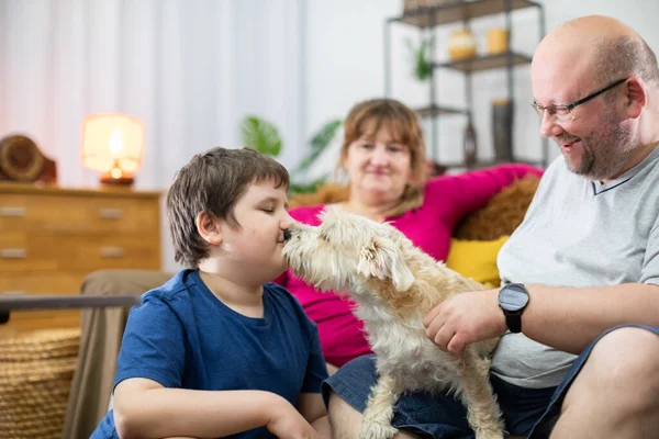 Um cão lambe o rosto de um menino que está sentado no sofá da sala de estar com seus pais. — Fotografia de Stock