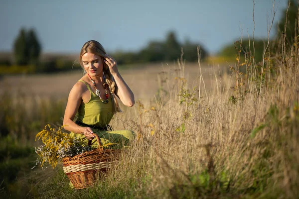 Una joven se agacha en un camino de tierra y mira las hierbas que ha cosechado y se arregla el cabello. —  Fotos de Stock