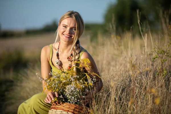 A young woman crouches on a dirt road and looks straight into the camera. A basket full of herbs. — Stock Photo, Image