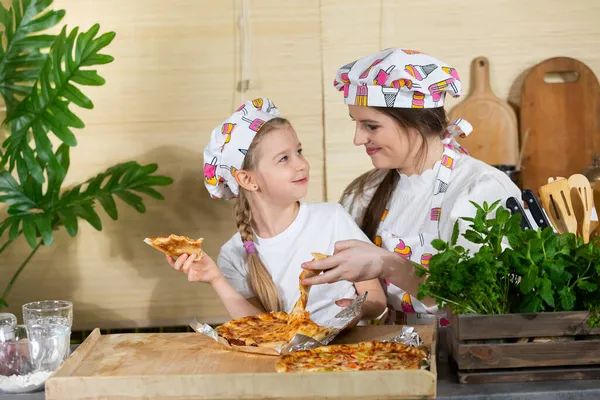 Пятилетняя дочь села на колени своих матерей, и они вместе едят пиццу, приготовленную и запечённую сами. После совместной работы на кухне, пришло время поесть вместе.. — стоковое фото