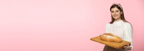 A young attractive saleswoman from a bakery shop holds a loaf of wheat flour bread on a wooden tray. The whole on a uniform light pink background. Panoramic frame. — Stock Photo, Image