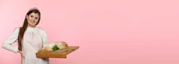 Panoramic frame. A young attractive saleswoman from a bakery shop holds a loaf of wheat flour bread and a pinch of fresh green herb leaves and rye ears on a wooden tray. The whole on a uniform light — Stock Photo, Image