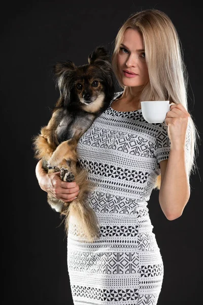 Genç bir kadın elinde bir köpek, diğerinde bir fincan kahve tutuyor. Uzun saçlı. Siyah arkaplan. — Stok fotoğraf
