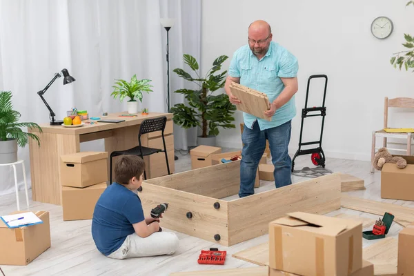 Filho senta-se e papai move as partes da estante em uma tigela para montagem. — Fotografia de Stock