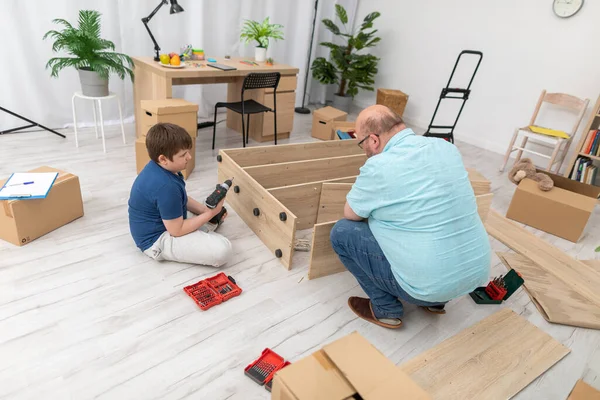 Filho senta-se e papai move as partes da estante em uma tigela para montagem. — Fotografia de Stock