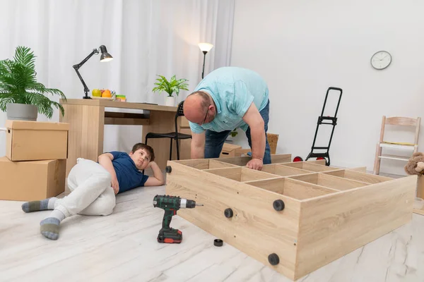 Papai laboriosamente monta móveis e filho apenas relógios. Montagem de mobiliário. — Fotografia de Stock