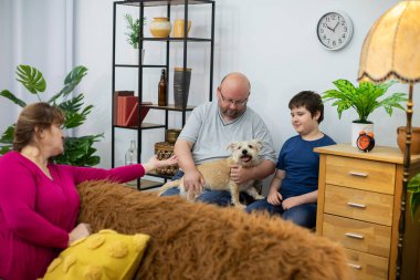 Bir baba ve oğul duvara yaslanmış sandalyelerde oturur ve tüylü bir köpeği kucağında tutarlar..