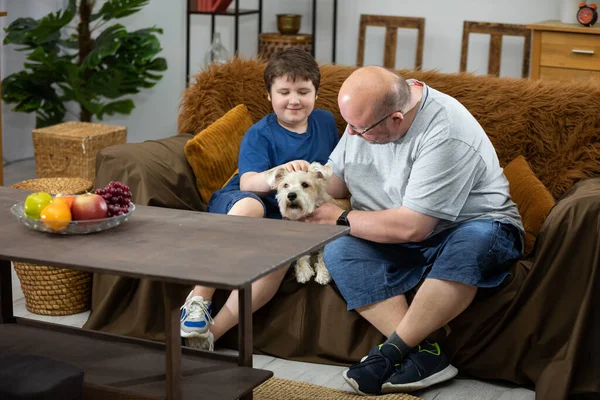 Um pai e um filho sentam-se num sofá e acariciam um cão castanho claro. — Fotografia de Stock