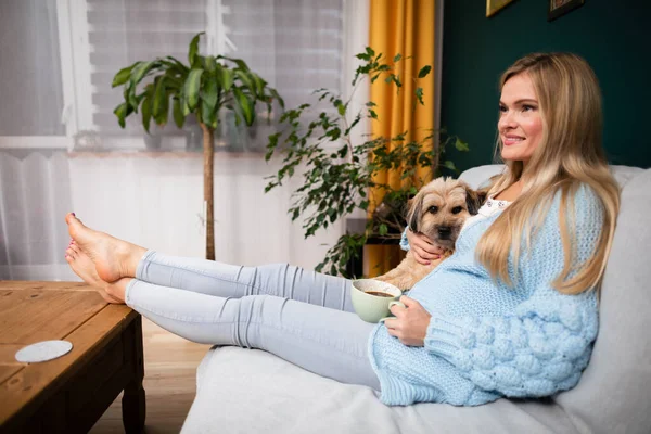 Uma menina está sentada em um sofá com uma xícara de café e um cão está deitado ao lado dela. — Fotografia de Stock