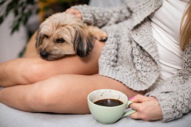 Bir kadın elinde kahve, yanında da çok kültürlü bir köpekle yatakta oturuyor..