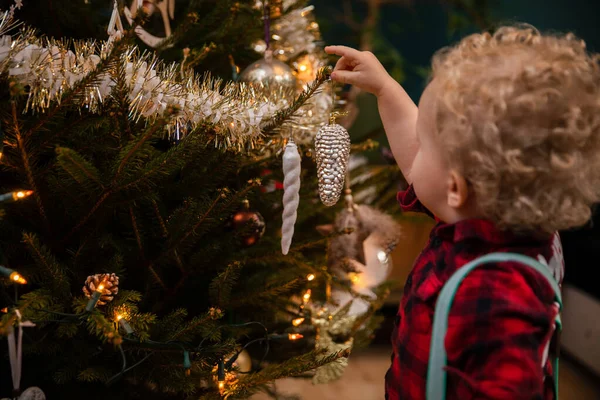 A two-year-old boy with blond curls hangs a bauble on a Christmas tree. — Stock Photo, Image