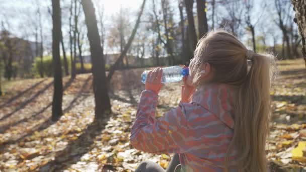 Ein Mädchen trinkt Wasser aus einer Flasche, während es in einem Park an einem Baum sitzt. — Stockvideo