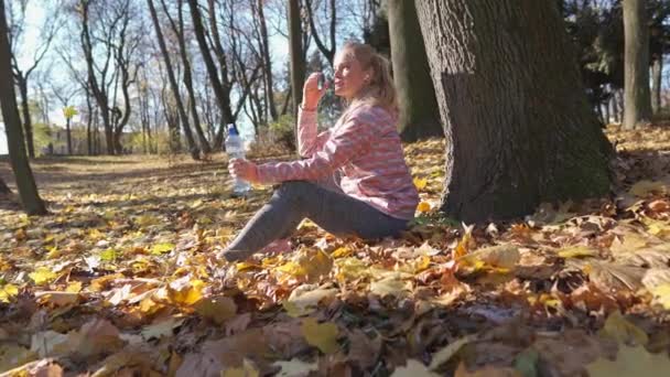 Bir kız sonbahar yapraklarının üzerine oturur ve şişeden su içer.. — Stok video