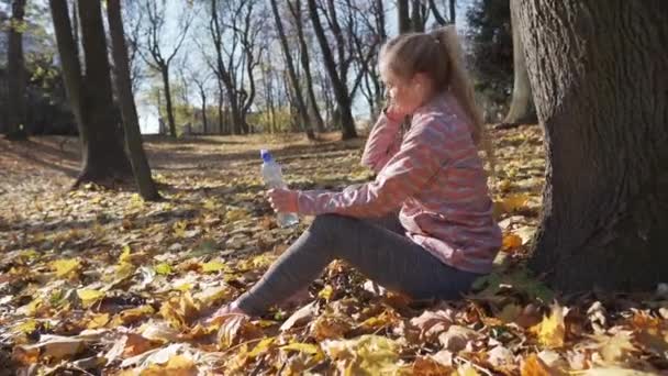 Een meisje zit op herfstbladeren bij een boom en drinkt water uit een fles.. — Stockvideo