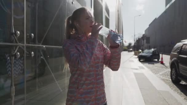 Egy nő sétál a városban, vizet iszik egy műanyag üvegből.. — Stock videók