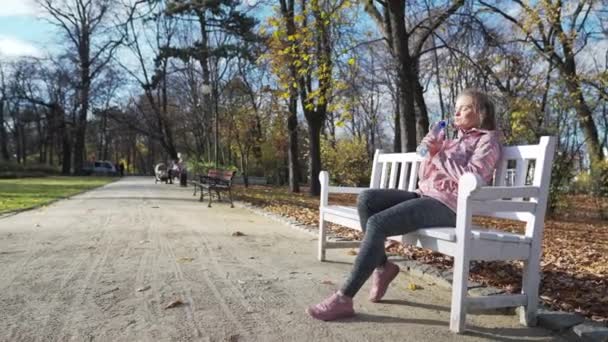 Una mujer atractiva se sienta en un banco y bebe agua de una botella de plástico. — Vídeo de stock