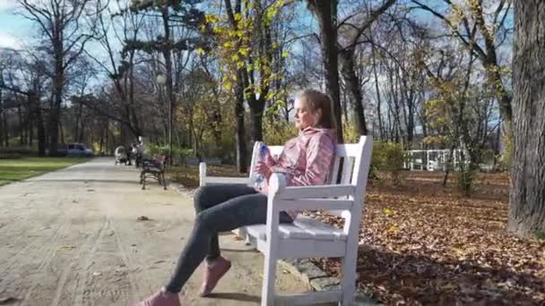 In a city park, a young woman is resting on a bench and drinking water. — стоковое видео