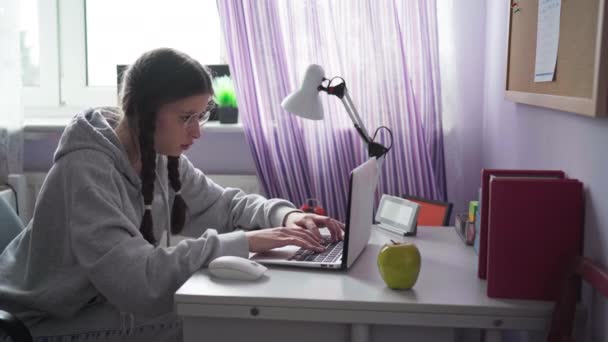 ノートパソコンのキーボードに十代の女の子が巧みにタイプします 眼鏡と二つの花嫁を持つ若い女の子は彼女の机に座って インターネットを使用しています — ストック動画