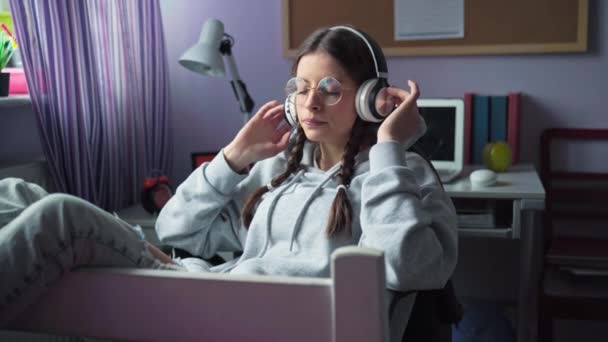 一个年轻的黑发姑娘的耳朵上戴着白色的耳机 在无线耳机上听音乐 戴眼镜和辫子的女孩 — 图库视频影像