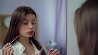 Genç bir kız dudaklarını rujla boyadı ve aynada şu an nasıl göründüğünü kontrol etti. Uzun ve dağınık saçlı genç bir esmer..