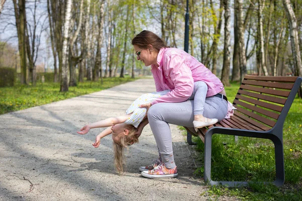 Outdoor gymnastics, mother and daughter, on a bench in the city park. Motor play with exercise of abdominal muscles in the child.