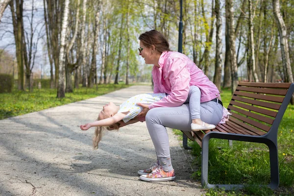 Outdoor gymnastics, mother and daughter, on a bench in the city park. Motor play with exercise of abdominal muscles in the child.