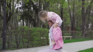 The daughter slowly gets off her mothers shoulders and sits on her lap and then from her lap walks and runs. A sunny spring day in the city park.