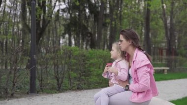 Bir anne ve kızı şehir parkında bir bankta oturuyorlar. Kız bir bidondan su içiyor ve ıslak dudaklarını koluyla siliyor. Güneşli bir bahar günü.