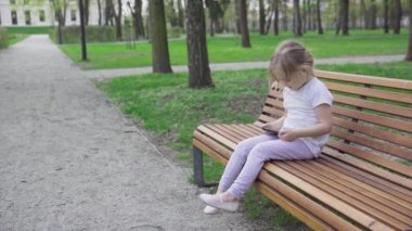 A four-year-old girl sits alone on a bench in a city park and watches cartoons on a smartphone. The child is holding a cell phone in her hand.