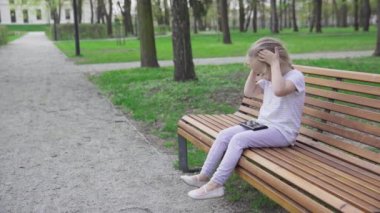 A four-year-old girl sits alone on a bench in a city park and watches cartoons on a smartphone. The child is holding a cell phone in her hand.