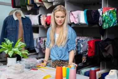 A blonde woman at a tailor shop is trying to choose the right thread color for her next blouse. A womens clothing designer.