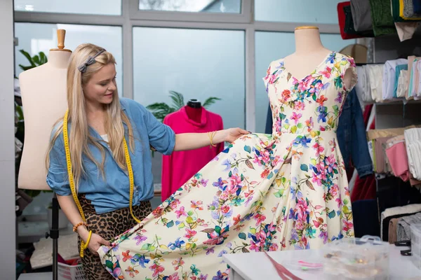 A clothing designer hangs a floral dress on a mannequin and checks how the whole thing looks. A blonde woman in the back of a tailors workshop.