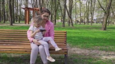 On a bench in a city park, a girl sits on her sitters lap and uses a smartphone. A sunny spring day outdoors.