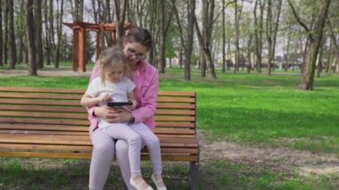 On a bench in a city park, a girl sits on her sitters lap and uses a smartphone. A sunny spring day outdoors.