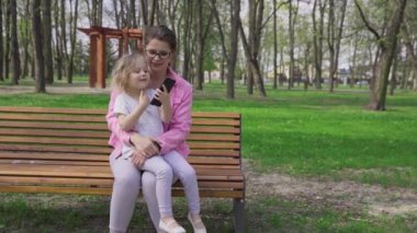 Relaxing on a bench in a city park with a smartphone in hand. A blonde and a brunette relax on a spring and sunny day.