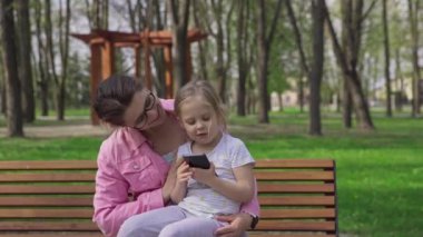 Tender parental kisses. Mother and daughter at leisure on a bench in a city park. A sunny spring day outdoors.