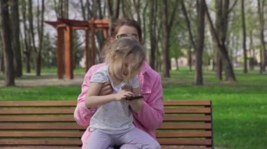 Relaxation and fun for mother and daughter on a sunny spring day. Spending time together in the city park. A four-year-old holds a smartphone in her hand.