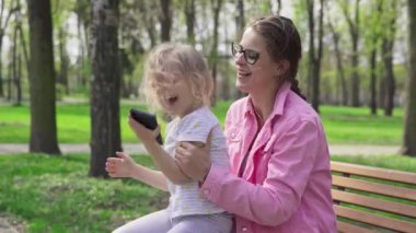 Joyful play while daughter sits on moms lap. A girl and her guardian laugh while sitting on a bench in a city park.