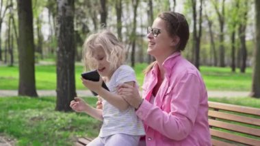 Joyful play while daughter sits on moms lap. A girl and her guardian laugh while sitting on a bench in a city park.