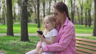 On a bench in a city park, a little girl sits on her mothers lap. The child busy all the time operating a smartphone. Shared moments in the fresh air.