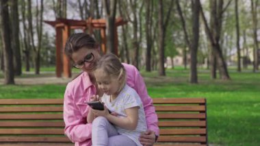 Fun to take a video selfie outdoors. A young mother and her four-year-old sit on a bench in a city park. Spring sunny day.