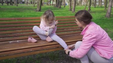 A mother helps her daughter remove a sock from her foot. A caregiver with a child in a city park. A girl with blonde hair sits on a park bench.