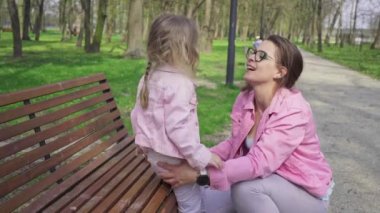 A daughter sits on a park bench and every so often comes down to kiss her mother. A girl with blonde hair and braided hair is playing with her mother.