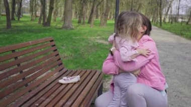 A daughter sits on a park bench and every so often comes down to kiss her mother. A girl with blonde hair and braided hair is playing with her mother.