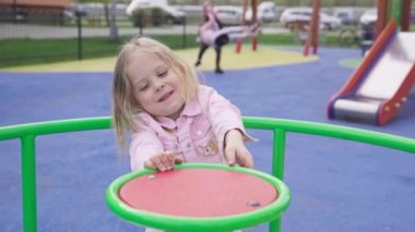 A fun playground game of spinning on a carousel. A little girl with blonde hair tied in a braid smiles and spins around.