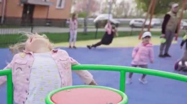 A fun playground game of spinning on a carousel. A little girl with blonde hair tied in a braid smiles and spins around.