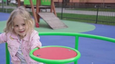 A girl spins by herself on a merry-go-round at a childrens playground. The little blonde girl with braided hair is laughing and happy.