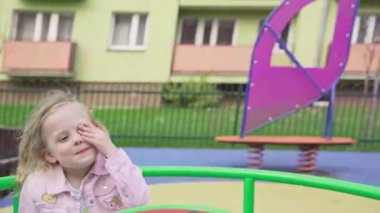 A girl spins by herself on a merry-go-round at a childrens playground. The little blonde girl with braided hair is laughing and happy.